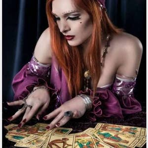 May include: A person with long red hair wearing a purple headscarf and a purple off-the-shoulder top with ruffled sleeves, adorned with silver armbands and rings, looking down at a spread of illustrated tarot cards on a dark textured surface.