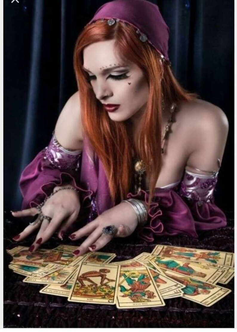 May include: A person with long red hair wearing a purple headscarf and a purple off-the-shoulder top with ruffled sleeves, adorned with silver armbands and rings, looking down at a spread of illustrated tarot cards on a dark textured surface.