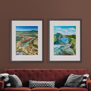 May include: Two framed landscape paintings with a red couch in the foreground. The paintings are both in black frames and feature colorful landscapes with water and trees.