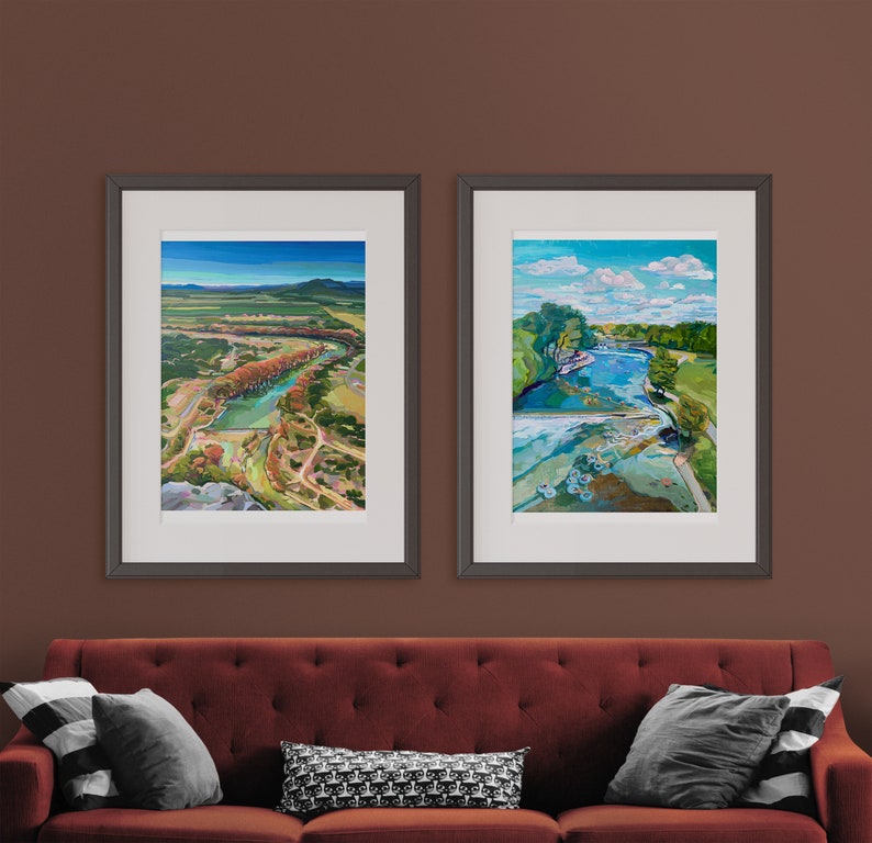 May include: Two framed landscape paintings with a red couch in the foreground. The paintings are both in black frames and feature colorful landscapes with water and trees.