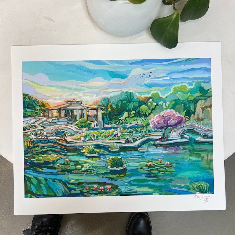 May include: A painting of a Japanese garden with a pond, bridge, and a small building. The pond is filled with lily pads and the water is a vibrant blue-green. The sky is a light blue with white clouds. The painting is signed "Craig Fuller 2022".
