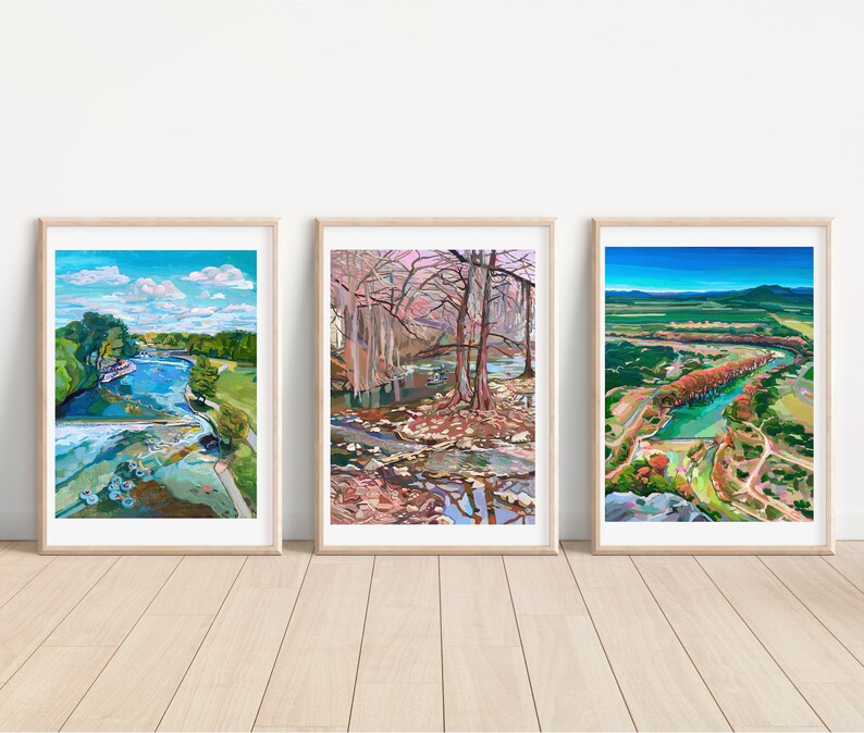 May include: Three framed landscape paintings of a river scene. The first painting shows a river with trees and a blue sky with clouds. The second painting shows a river with trees and a pink sky with clouds. The third painting shows a river with trees and a blue sky with mountains in the distance.