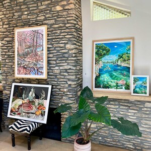K&ouml;nnte beinhalten: Ein Wohnzimmer mit gerahmten Kunstdrucken und einer Topfpflanze. Die Kunst umfasst eine Landschaft mit einem Fluss, eine Szene mit schwimmenden Menschen und ein Stillleben mit Essen. Die Pflanze hat gro&szlig;e, gr&uuml;ne Bl&auml;tter und einen rosa-wei&szlig;en Topf.