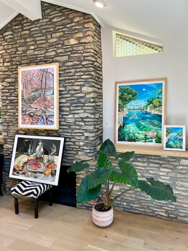 K&ouml;nnte beinhalten: Ein Wohnzimmer mit gerahmten Kunstdrucken und einer Topfpflanze. Die Kunst umfasst eine Landschaft mit einem Fluss, eine Szene mit schwimmenden Menschen und ein Stillleben mit Essen. Die Pflanze hat gro&szlig;e, gr&uuml;ne Bl&auml;tter und einen rosa-wei&szlig;en Topf.