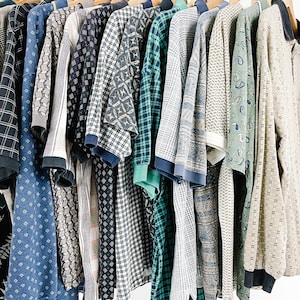 May include: A collection of long-sleeved shirts hanging on wooden hangers, featuring a variety of patterns including plaids, geometric shapes, and paisley prints in shades of blue, green, beige, and black, all arranged in a row on a rack.