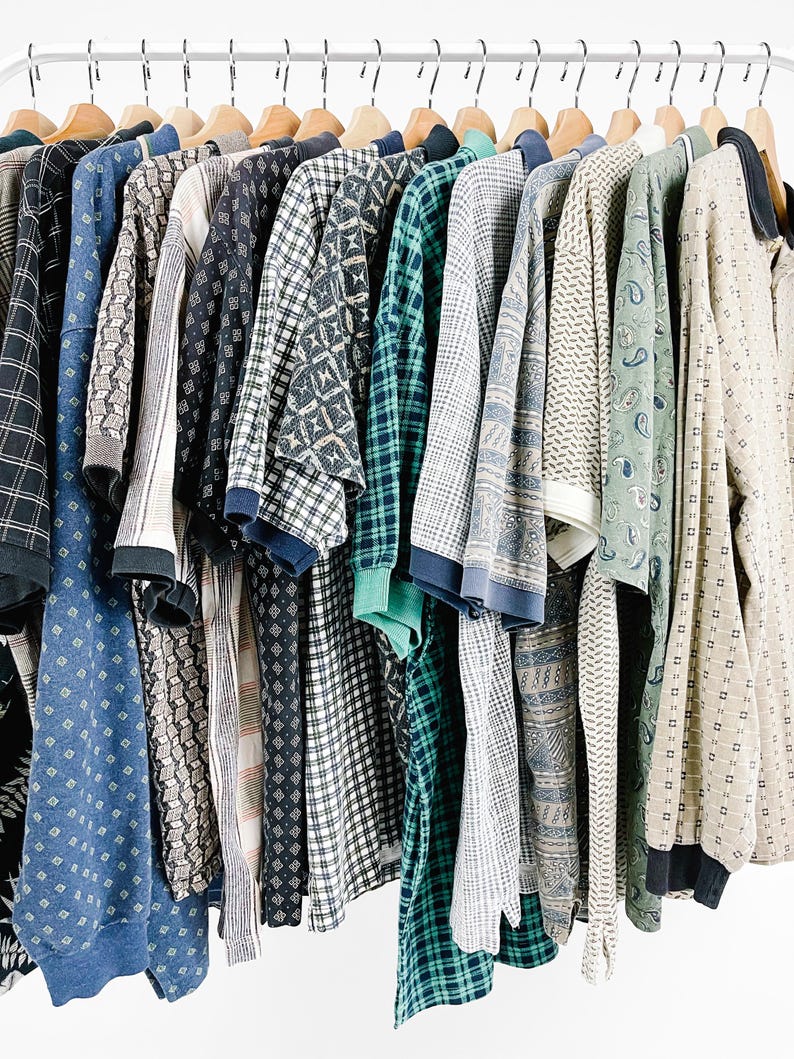May include: A collection of long-sleeved shirts hanging on wooden hangers, featuring a variety of patterns including plaids, geometric shapes, and paisley prints in shades of blue, green, beige, and black, all arranged in a row on a rack.