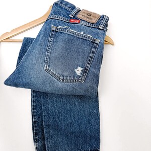 Puede incluir: Vaqueros Wrangler azules con un aspecto desgastado, con un parche desgastado en el bolsillo trasero. Los vaqueros est&aacute;n colgados de una percha de madera. El logotipo de Wrangler es visible en el bolsillo trasero.