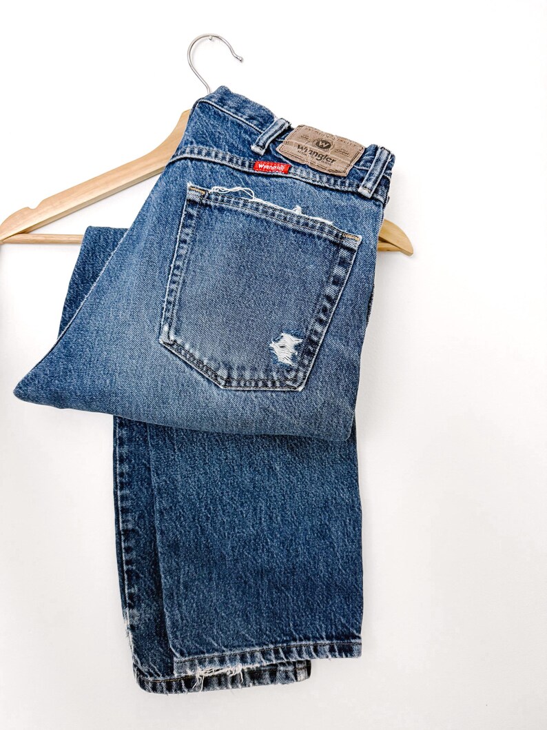 Puede incluir: Vaqueros Wrangler azules con un aspecto desgastado, con un parche desgastado en el bolsillo trasero. Los vaqueros est&aacute;n colgados de una percha de madera. El logotipo de Wrangler es visible en el bolsillo trasero.