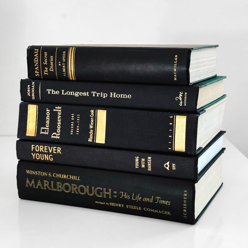 May include: A stack of five hardcover books with black covers and gold lettering. Titles include "Spandau: The Secret Diaries," "The Longest Trip Home," and "Eleanor Roosevelt." The books are arranged on a white surface.