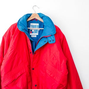 May include: A bright red jacket with a contrasting blue inner collar and lining. The garment features a front zipper closure, black snap buttons, and a label on the inside neck that reads Trader Bay with a small red, white, and blue square logo.