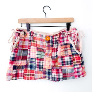 May include: A plaid mini skirt featuring a patchwork design with various shades of red, pink, yellow, blue, and white. The skirt includes a bright orange button at the center waist and light beige drawstring ties on both sides with metal grommets.