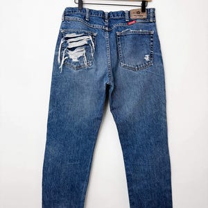 Puede incluir: Vaqueros azules de mezclilla con detalles desgastados, incluyendo secciones rasgadas en los bolsillos traseros. Los vaqueros est&aacute;n colgados en una percha de madera. La etiqueta de la marca Wrangler es visible.