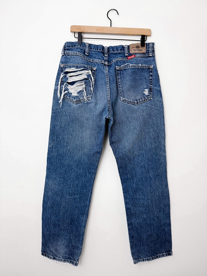 Puede incluir: Vaqueros azules de mezclilla con detalles desgastados, incluyendo secciones rasgadas en los bolsillos traseros. Los vaqueros est&aacute;n colgados en una percha de madera. La etiqueta de la marca Wrangler es visible.