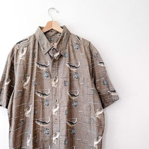 May include: A short-sleeved button-down shirt in a neutral brown color, adorned with a repeating pattern of fish and fishing-related imagery. The shirt is hanging on a wooden hanger against a white background.