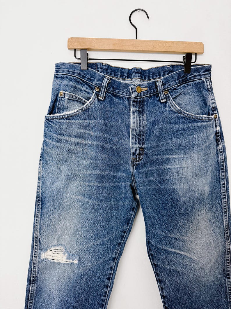 Puede incluir: Vaqueros azules de mezclilla con una zona de rodilla desgastada, colgados de una percha de madera. Los vaqueros tienen un dise&ntilde;o cl&aacute;sico de cinco bolsillos, cierre de bot&oacute;n y cremallera, y un lavado descolorido. Los vaqueros son de un azul medio.