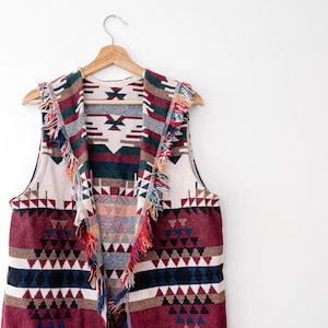 May include: A sleeveless vest featuring a woven pattern in shades of burgundy, cream, dark green, and navy blue. The garment has a wide collar with multi-colored fringe trim along the edges and a geometric design throughout the fabric.