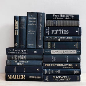 May include: A stack of navy blue hardcover books with gold lettering. Titles include "Josh Logan", "The Columbia History of the World", and "The Fifties". The books are arranged on a white surface.