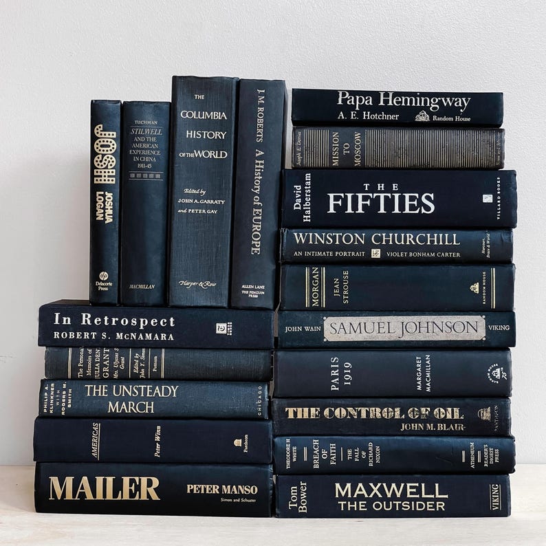May include: A stack of navy blue hardcover books with gold lettering. Titles include "Josh Logan", "The Columbia History of the World", and "The Fifties". The books are arranged on a white surface.