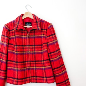 May include: A red plaid jacket with a fuzzy texture, featuring a full-length front zipper, a pointed collar, and long sleeves. The fabric displays a pattern of intersecting black and purple lines. A black label with the text REQUIREMENTS is visible.