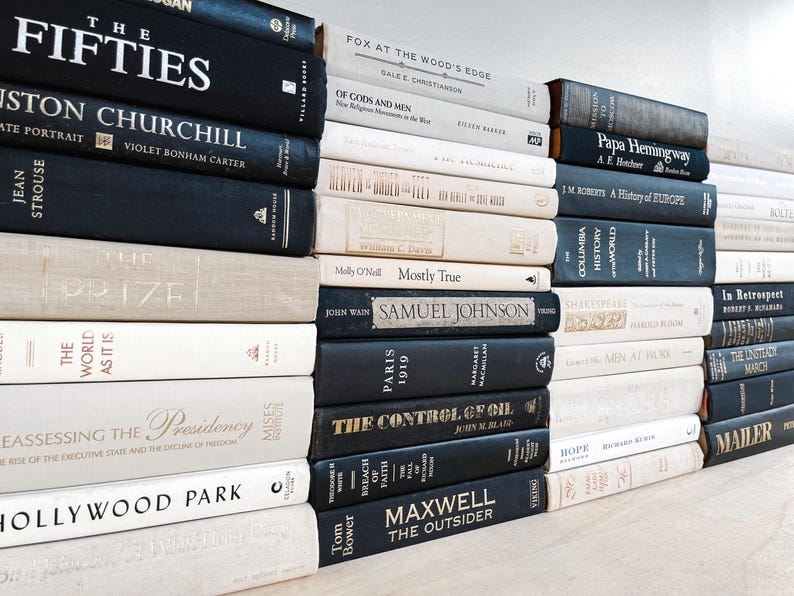 May include: A collection of hardcover books in various sizes and colors. The spines display titles like "The Fifties," "Hollywood Park," and "The Control of Oil." The books are stacked on a light-colored surface, creating a visually appealing arrangement.