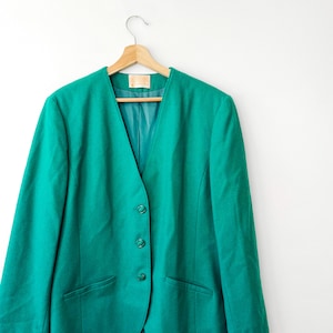 May include: A teal blazer with a V-neck design, three matching buttons, and two front pockets. The blazer is hanging on a wooden hanger against a white background. The blazer has a teal lining.