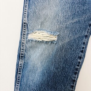Puede incluir: Primer plano de unos vaqueros azules con una rodilla desgastada. Los vaqueros tienen un lavado descolorido y costuras visibles a lo largo de las costuras. El &aacute;rea desgastada revela la tela subyacente.