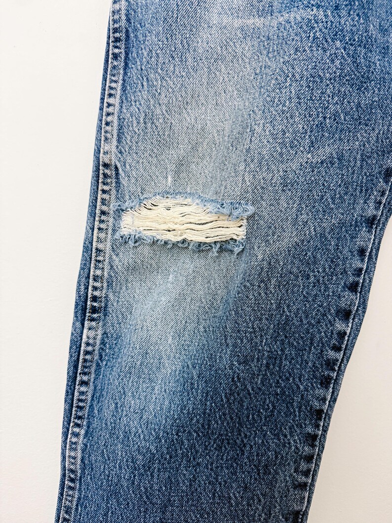 Puede incluir: Primer plano de unos vaqueros azules con una rodilla desgastada. Los vaqueros tienen un lavado descolorido y costuras visibles a lo largo de las costuras. El &aacute;rea desgastada revela la tela subyacente.
