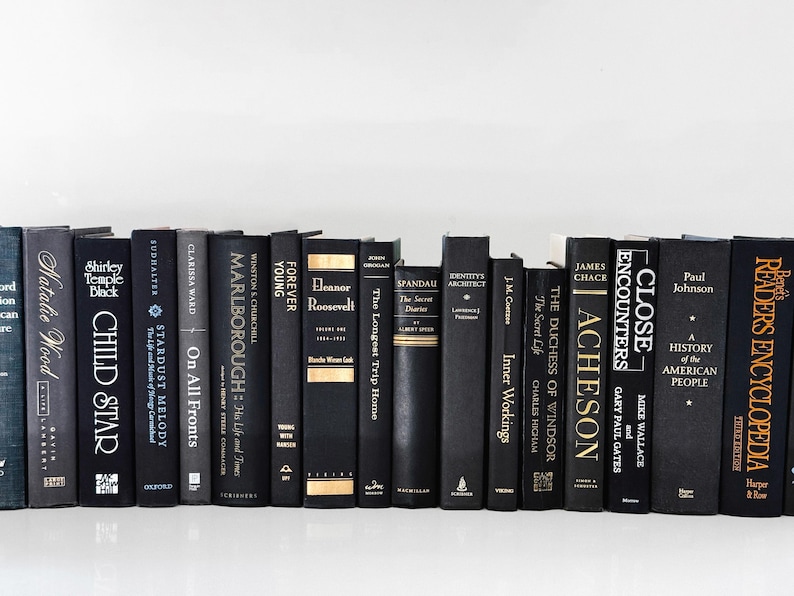 May include: A collection of hardcover books with black spines and various titles. The books are arranged in a row, with titles and author names visible. The spines have gold or white lettering. The books are on a white surface.