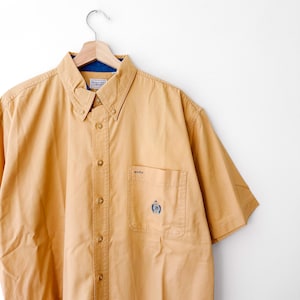 May include: A short-sleeved button-down shirt in a solid tan color. The shirt features a button-down collar, a front button placket, and a single chest pocket with a small embroidered logo. The inner collar lining is a contrasting dark blue fabric.