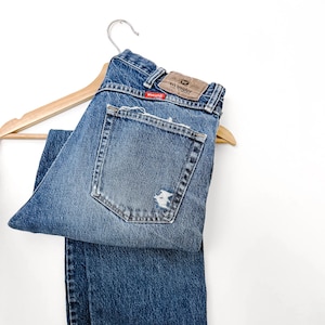 Puede incluir: Vaqueros azules de mezclilla con un aspecto desgastado, colgados de una percha de madera. Los vaqueros tienen un aspecto descolorido con una secci&oacute;n rasgada en el bolsillo trasero. La marca "Wrangler" es visible en un parche de cuero.