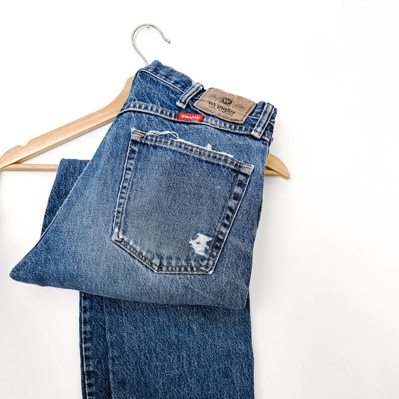 Puede incluir: Vaqueros azules de mezclilla con un aspecto desgastado, colgados de una percha de madera. Los vaqueros tienen un aspecto descolorido con una secci&oacute;n rasgada en el bolsillo trasero. La marca "Wrangler" es visible en un parche de cuero.
