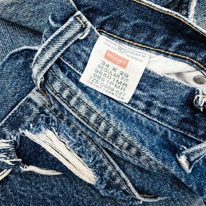 Puede incluir: Primer plano de unos vaqueros de mezclilla azules con una etiqueta Wrangler. Los vaqueros son de corte regular, talla 34 x 29. La mezclilla tiene un aspecto desgastado con bordes deshilachados. La etiqueta indica que los vaqueros son 100% algod&oacute;n.