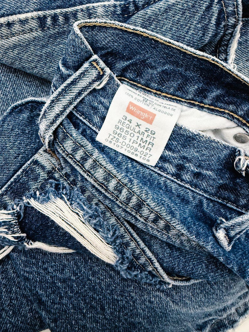 Puede incluir: Primer plano de unos vaqueros de mezclilla azules con una etiqueta Wrangler. Los vaqueros son de corte regular, talla 34 x 29. La mezclilla tiene un aspecto desgastado con bordes deshilachados. La etiqueta indica que los vaqueros son 100% algod&oacute;n.
