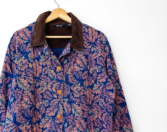 Puede incluir: Una camisa de manga larga con botones y un estampado de hojas azules y rosas. La prenda tiene un cuello de pana marrón oscuro y botones redondos de color marrón claro en la parte delantera. Se muestra en una percha de madera.