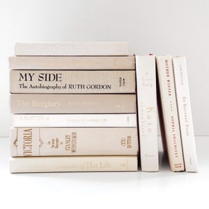 May include: A collection of hardcover books in neutral beige and cream tones. The stack includes titles such as My Side, The Burglary, Victoria, Kate Remembered, Mother Winter, and On Borrowed Words, with visible black and metallic gold lettering.