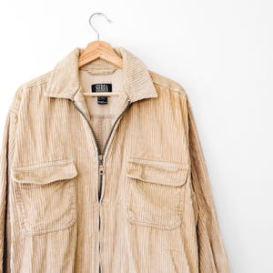 May include: A tan corduroy jacket with a full-length front zipper, a pointed collar, and two chest pockets with flaps. The interior label features the text SIERRA. The jacket is displayed on a wooden hanger against a plain white background.