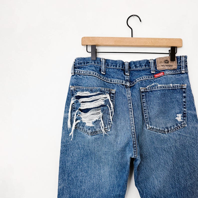 Puede incluir: Vaqueros Wrangler de mezclilla azul con detalles desgastados. Los vaqueros est&aacute;n colgados de una percha de madera. Los bolsillos traseros y la etiqueta Wrangler son visibles. Los vaqueros tienen detalles rasgados en la espalda.