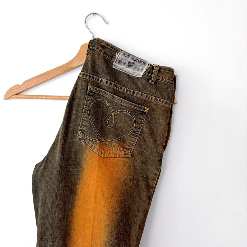 Puede incluir: Vaqueros de mezclilla marrones y naranjas con un dise&ntilde;o en forma de coraz&oacute;n en el bolsillo trasero. Los vaqueros cuelgan de una percha de madera. La etiqueta dice "JOE BOXER".