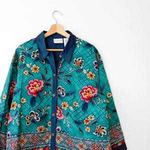 May include: A teal quilted button-down shirt with a floral pattern of pink, yellow, and orange flowers. The shirt has a navy blue collar and trim. The shirt is hanging on a wooden hanger.