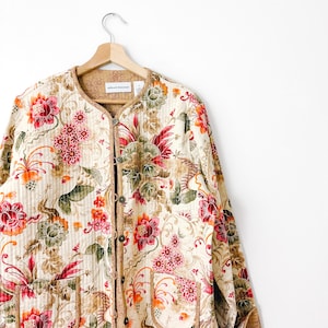 May include: Cream-colored quilted jacket with a floral pattern in red, pink, orange, and green. The jacket has a round collar, button closure, and is hanging on a wooden hanger. The floral design is intricate and covers the entire jacket.