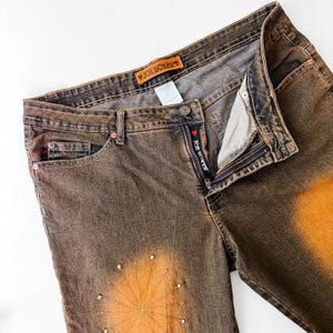 Puede incluir: Vaqueros de mezclilla marrones con un dise&ntilde;o descolorido y un patr&oacute;n decorativo en la parte delantera. Los vaqueros tienen una bragueta con cremallera y la marca "JOE BOXER" es visible en una etiqueta y la cremallera. Los vaqueros est&aacute;n planos sobre una superficie blanca.