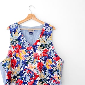 May include: A sleeveless vest featuring a vibrant floral pattern with red, yellow, and blue flowers on a white background. The garment has a V-neckline, a front button closure with white buttons, and light blue side panels. A Ruff Hewn label is visible.