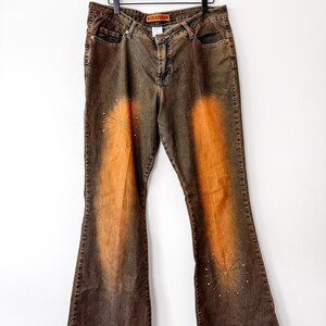 Puede incluir: Vaqueros acampanados marrones y naranjas con un dise&ntilde;o descolorido y adornos de estrellas. Los vaqueros cuelgan de una percha de madera. El nombre de la marca "ICE BOXERS" es visible en la cintura.