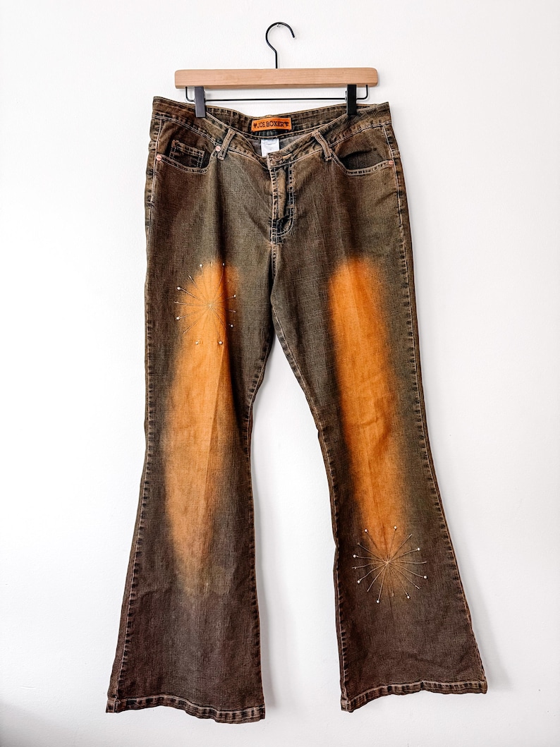 Puede incluir: Vaqueros acampanados marrones y naranjas con un dise&ntilde;o descolorido y adornos de estrellas. Los vaqueros cuelgan de una percha de madera. El nombre de la marca "ICE BOXERS" es visible en la cintura.