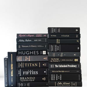 May include: A stack of hardcover books with black covers and gold or white lettering. Titles include "Titan," "The Fifties," and "John Adams." The books are arranged in a neat pile against a white background, creating a minimalist aesthetic.