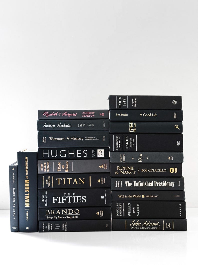 May include: A stack of hardcover books with black covers and gold or white lettering. Titles include "Titan," "The Fifties," and "John Adams." The books are arranged in a neat pile against a white background, creating a minimalist aesthetic.