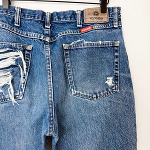 Puede incluir: Primer plano de vaqueros de mezclilla azul desgastados con detalles rasgados en los bolsillos traseros y la pierna izquierda. La etiqueta de la marca Wrangler es visible en la cintura trasera. Los vaqueros cuelgan de una percha de madera.