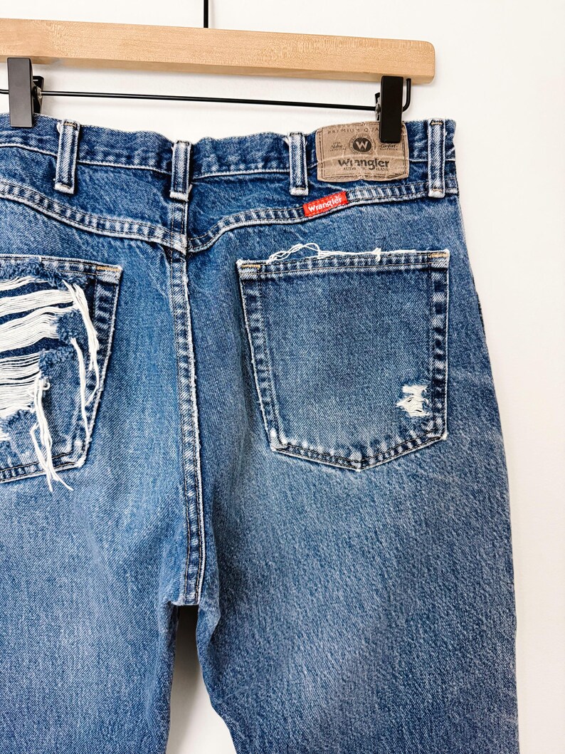Puede incluir: Primer plano de vaqueros de mezclilla azul desgastados con detalles rasgados en los bolsillos traseros y la pierna izquierda. La etiqueta de la marca Wrangler es visible en la cintura trasera. Los vaqueros cuelgan de una percha de madera.