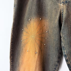 Puede incluir: Primer plano de unos vaqueros de mezclilla con un dise&ntilde;o de sol. Los vaqueros tienen un lavado azul descolorido con un degradado naranja-marr&oacute;n. El sol est&aacute; bordado con hilo dorado y adornado con peque&ntilde;as cuentas blancas.