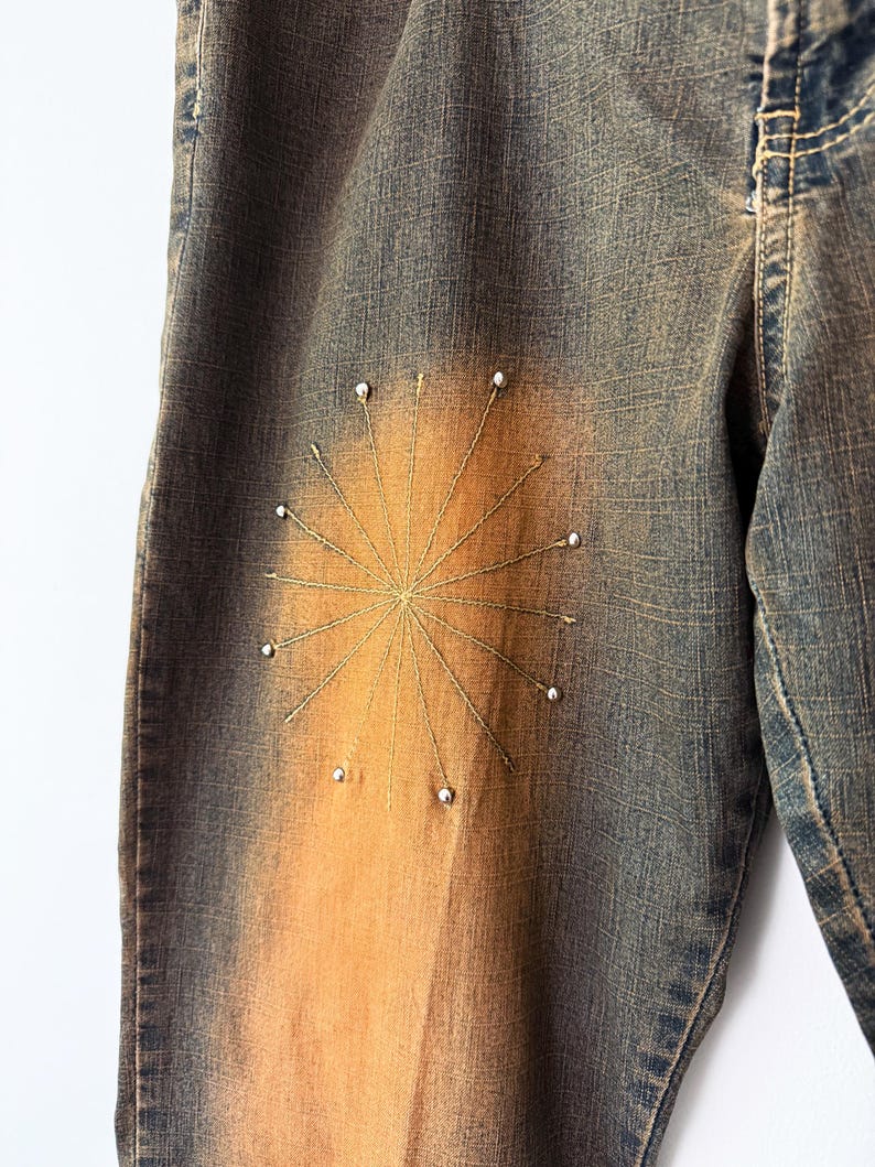 Puede incluir: Primer plano de unos vaqueros de mezclilla con un dise&ntilde;o de sol. Los vaqueros tienen un lavado azul descolorido con un degradado naranja-marr&oacute;n. El sol est&aacute; bordado con hilo dorado y adornado con peque&ntilde;as cuentas blancas.